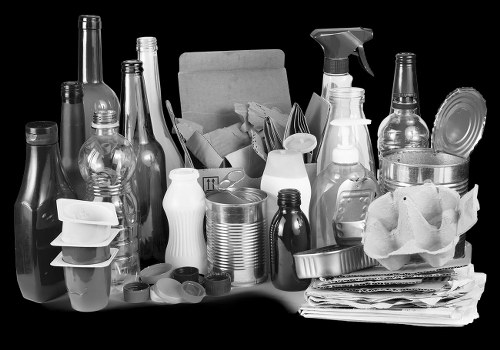 A collection of various discarded household items and packaging materials, including plastic bottles, glass bottles, aluminium cans, paper and cardboard, and miscellaneous waste, all stacked and spread across a surface. The arrangement appears cluttered with some bottles standing upright, while others are leaning or stacked. There are crumpled paper and newspaper on the right side, and the background is plain and dark, emphasizing the waste items. The scene is indicative of rubbish accumulation suitable for removal services, such as those provided by Office Clearance Hornsey, with items stored in an outdoor or indoor workspace, ready for disposal or collection.