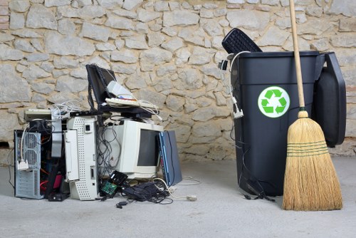 A pile of discarded electronic waste including old computer towers, monitors, and various cables positioned on a concrete surface next to a black recycling bin with a green recycling symbol, which contains a few black plastic items and a wooden-handled broom leaning against it. The scene is set outdoors, with a textured beige stone wall in the background, suggesting a residential or commercial area where rubbish removal services may be required in Hornsey or surrounding locations. The electronic waste appears to be in a disorganized state, awaiting collection or disposal by professional rubbish removal services such as those offered by Office Clearance Hornsey. The environment is well-lit, emphasizing the contrast between the rough stone wall and the cluttered foreground, illustrating typical waste items targeted for removal and recycling within the local waste management context.