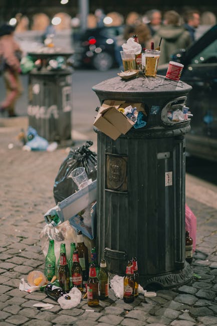 A black cylindrical metal rubbish bin with a domed lid stands on a cobbled pavement in an urban setting. The lid is partially obscured by various waste items placed on top, including crumpled paper cu