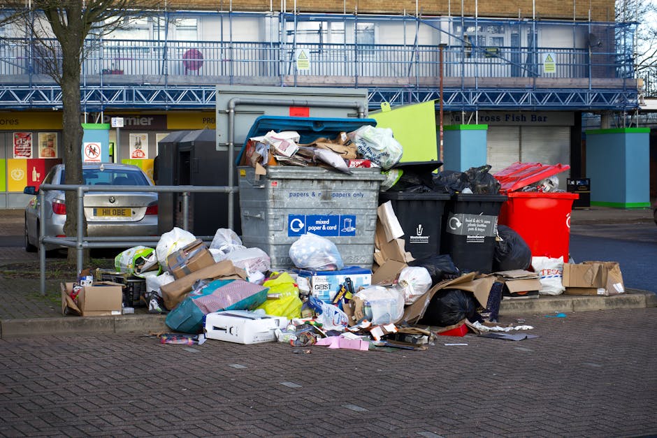 A black cylindrical metal rubbish bin with a domed lid stands on a cobbled pavement in an urban setting. The lid is partially obscured by various waste items placed on top, including crumpled paper cu