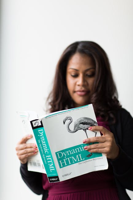 A woman with medium-length dark brown hair is holding a book titled 'Dynamic HTML' by O'Reilly in front of a plain white wall. She is dressed in a dark blazer over a maroon top. The book cover feature