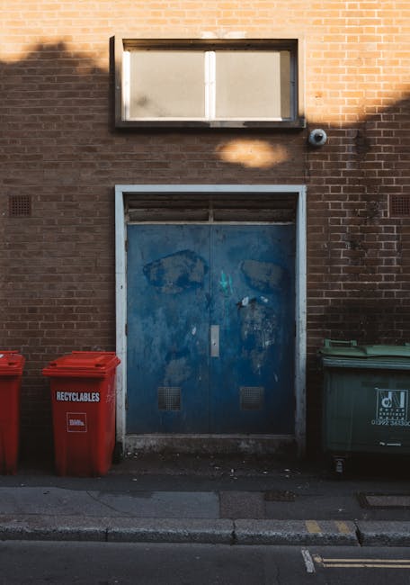 The image displays a section of a brick building exterior featuring a large, weathered blue metal door with visible scratches, rust, and peeling paint. Above the door, there is a rectangular window wi